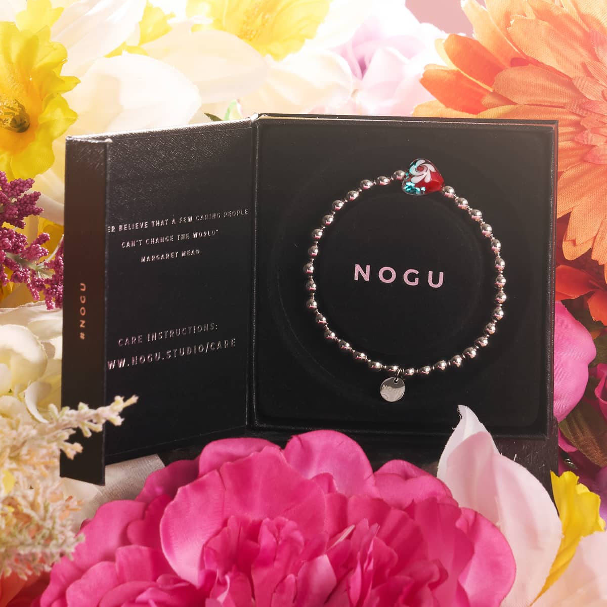 Crimson Swirl Glowing Heart Essence Bracelet with silver beads and red and white heart charm in black NOGU box surrounded by pink and yellow flowers